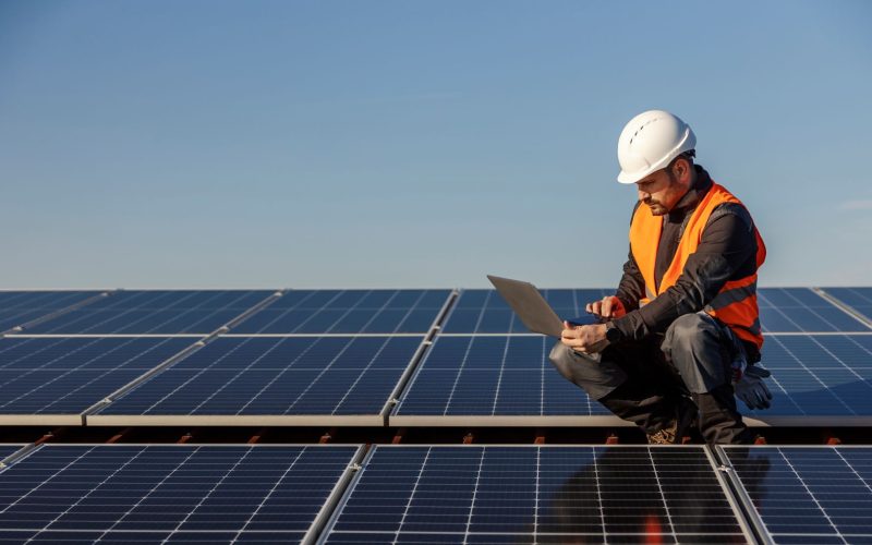 Engineer reviews data on his laptop while standing next to large solar panels.